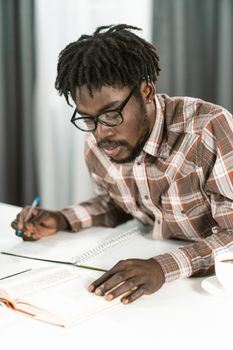 African American student read book and makes notes in his copybook doing his homework. Portrait young man in glasses study at home or library. Educational concept.