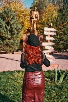 Stylish young woman stands with her back against the background of a monument in the park. A young woman with red hair wearing a beret and a leather pencil skirt.