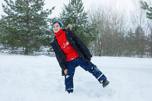A cheerful teenager in a dark jacket and a red sweater stands in a winter park on the snow