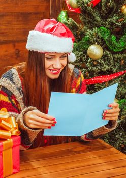 Pretty White Woman in a Casual Warm Sweater and Santa Hat Reading a Christmas Card Next to the Christmas Tree with Several Glitter Balls . Young Lady is Enjoys Reading Christmas Greetings. Blank Cover. Copy Space. Close-up. Wood Background. High quality photo