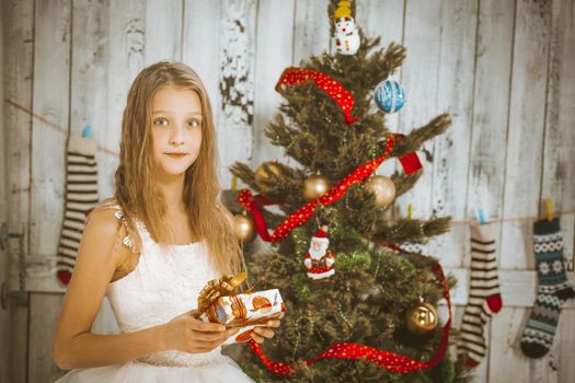 caucasian girl model with long wavy blond hair in a white Fluffy dress with a gift in her hands stands by a Christmas tree decorated with balls. Girl looks at the camera in the Christmas New Year studio. In the background are Christmas socks with ornaments on the wall. Close-up. High quality photo