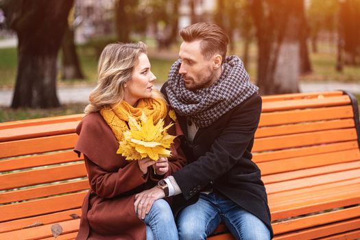 happy loving couple embracing sitting on the bench romantic hugged in park wearing coats and scarfs Collecting a bouquet of fallen leaves. Love story concept. Tinted image.