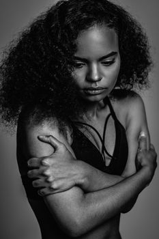 Black and white image. Standing sideways tender African American girl dressed in black top isolated on grey background. Human emotions, facial expression concept.