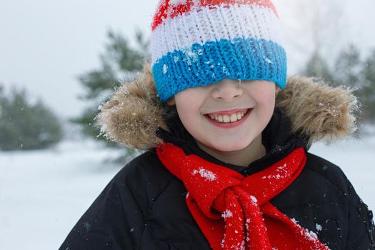 Close up portrait of a happy boy in bright winter clothes and a red scarf stands in a striped hat pulled down over his eyes, smiling, in a winter park with pine trees