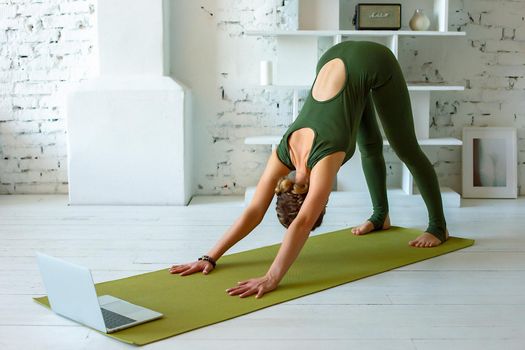 A slender woman in a green sports jumpsuit performs the -dog face down, yoga exercise at home, in a light interior, in front of a laptop