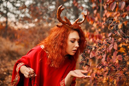 Woman in long red dress with deer horns in autumn forest. Beautiful mystery gothic creature in long red cloak with deer horns in autumn forest. Dark fantasy creature elf in magic wood