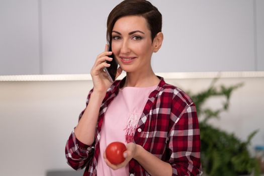Talking on the phone with tomato in the hand young pretty housewife with a bob hairstyle prepares food in the kitchen. Healthy food at home. Healthy food leaving - vegan concept.