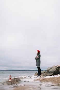 Winter portrait of young woman in a coat and red hat holding decorative lighthouse and standing at the shore of frozen sea. winter, travel, sea background. windy weather, amazing ice seaside
