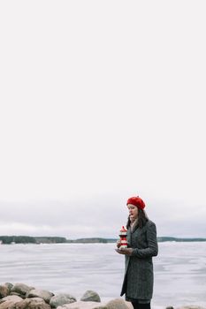 Winter portrait of young woman in a coat and red hat holding decorative lighthouse and standing at the shore of frozen sea. winter, travel, sea background. windy weather, amazing ice seaside