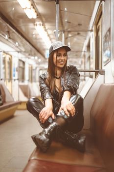 Smiling beautiful brunette female in modern latex clothes sits with folded legs on the seat in the empty metro car. Nice woman inside the night underground. Close-up Portrait. Toned picture. High quality photo