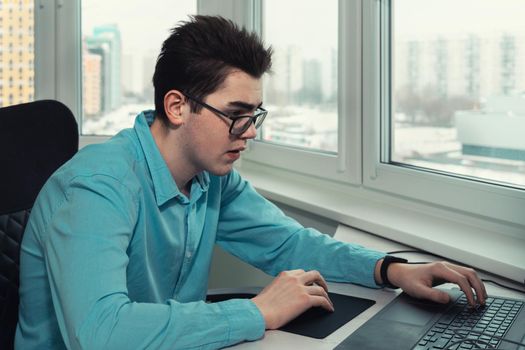 young employee with glasses looking at laptop screen and typing.