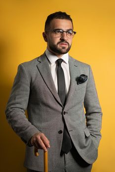 Successful handsome english style young man wearing grey suit standing holding a cane or umbrella in his hand isolated on yellow background.