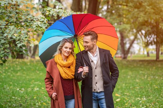 A beautiful young couple is walking in the park under a rainbow colored umbrella. A beautiful girl walks through the autumn park arm in arm with a guy in rainy weather.