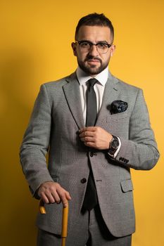 English style handsome young man wearing grey suit with one hand lifted standing with cane or umbrella in his hand isolated on yellow background.