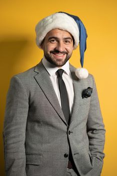 Young handsome bearded smiling Santa man wearing blue Santa Claus hat and suit, looking at camera isolated on yellow background. Positive emotions, facial expressions, feelings seasonal holidays.