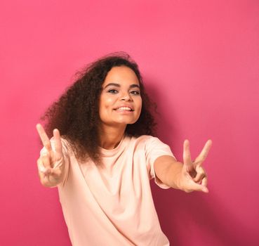 Happy sending PEACE gesture with two hands beautiful young African American woman looking positively at camera wearing peachy t-shirt isolated on pink background. Beauty concept. Square crop.