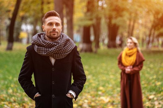 Man stands in a coat and a scarf in the park waiting for his girlfriend or wife who stands behind him or looks like him from the back. Young couple outdoors in a park on a beautiful autumn day.