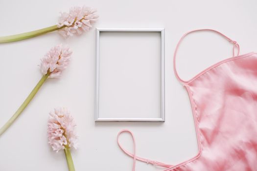 Blank photo frame, silk lingerie and pink flowers on white background. Styled feminine flat lay, top view, copy space. Creative concept, empty greeting card. 
