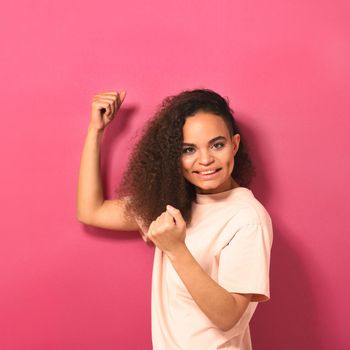 Happy beautiful young African American woman looking positively at camera wearing peachy t-shirt showing her muscles isolated on pink background. Beauty concept. Square crop.