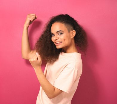 Happy and excited expressing winning gesture young African American girl looking positively at camera wearing peachy t-shirt isolated on pink background. Beauty concept. Square crop.