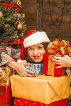 Lovely Young Woman in a Red Santa Hat Holding a Dozen of Christmas Multi Colored Gifts. Decorated Christmas Tree, Electric Garland on the Wall, Lights. Close-up. Wood Background. High quality photo