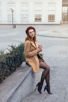 Beautiful girl in an autumn coat and beige sits on a marbled flower beds waiting for her date or girlfriends. Toned photo.