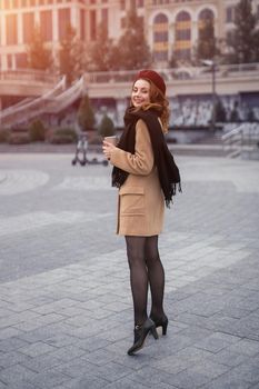 French styled young woman looking to camera turned head from back holding a coffee mug on the street. Portrait of stylish young woman wearing autumn coat and red beret outdoors. Tinted image.