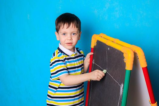 The boy draws on the blackboard with a chalk . A boy on a blue background. An article about children's leisure. Drawing board