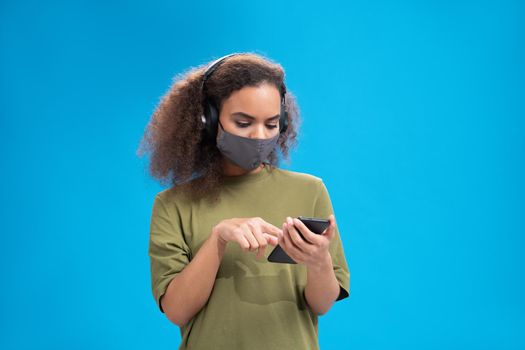 Dumb girl speaks on video call on her smartphone wearing headphones in olive t-shirt in reusable face mask, to prevent others from corona COVID-19 infection isolated on blue background.