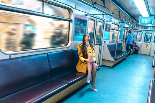 stylish lady in a sexy yellow coat and nude shoes sits on a seat with crossed legs in an empty subway car. Blurred background. medium shot. High quality photo