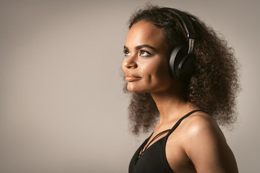 Pretty hipster girl African-American listening music in black earphones wearing black top isolated on grey background, emotionally move. Concept of emotions, facial expression.