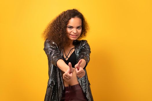 Positive beautiful ethnic girl with afro hair showing thumbs up smile looking at camera wearing shiny black jacket. Happy young girl standing on yellow background. Studio shot.