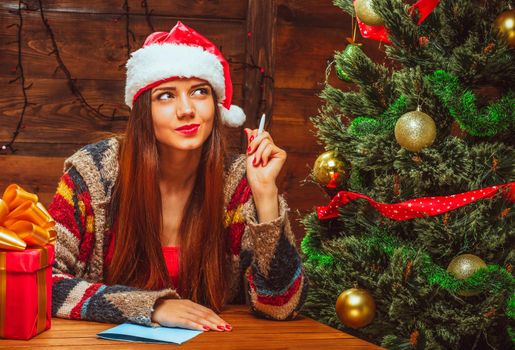 Charming Woman in a Red Hat with White Fur Sits at a Table near a Decorated Christmas Tree and Thinks what to Write to Santa. Close-up. High quality photo