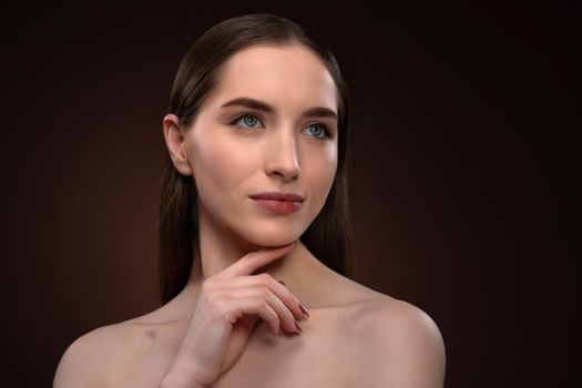 No make up portrait of a beautiful woman with a long hair and bare shoulders. Charming brunette model girl posing on camera touching her chin with a finger isolated on black background.