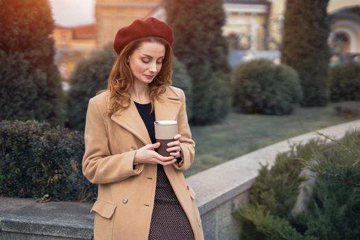 Portrait of stylish young woman wearing autumn coat and red beret outdoors. Pensive beautiful young woman holding a cup of coffee on the street. Female fashion concept. Autumn accessories.