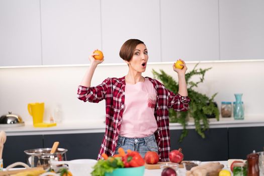 Excited woman with citrus fruits in hands standing in the modern kitchen. Woman in red plaid shirt making fresh citrus juices. Young female with short hair making organic drinks. Healthy life concept.