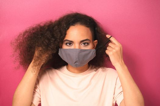 Taking off putting on reusable face mask African American girl in peachy t-shirt, to prevent others from corona COVID-19 and SARS cov 2 infection isolated on pink background.