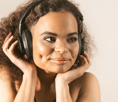 Young African-American girl listening music in headphones wearing black top isolated on grey background, emotionally move. Concept of emotions, facial expression. Square cropped.