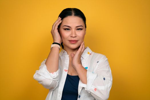 Cute mature Asian woman in a casual posing posh look on camera touching her face with hands. isolated on yellow background. Human emotions, facial expression concept.