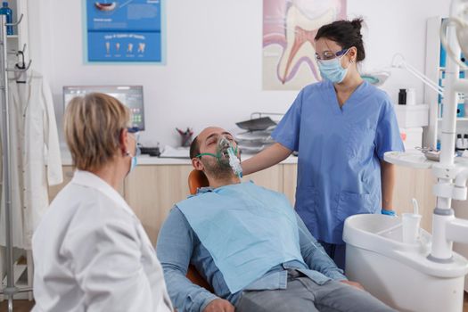 Orthodontist team putting oxygen mask on man patient before starting dental surgery during stomatological consultation in dentistry office room. Specialist assistant working at caries treatment