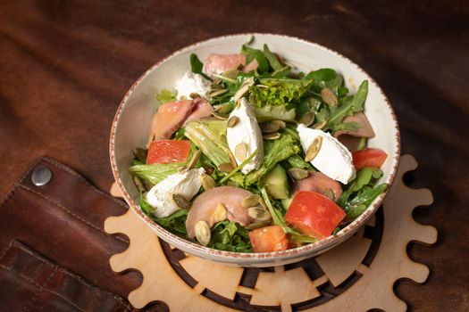 Bowl of fresh vegetable salad with slices of salmon, mozzarella cheese stands on a table covered with a leather rag and a decorative detail of a simple mechanism. Restaurant concept.