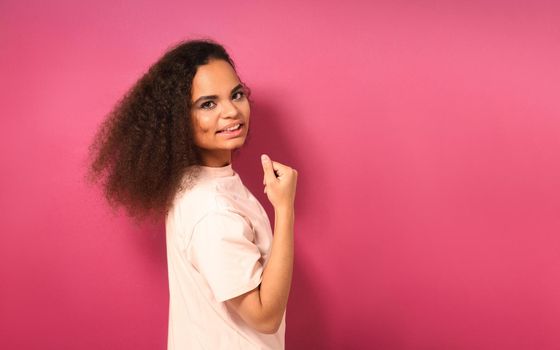 Beautiful young African American girl half turned looking positively at camera wearing peachy t-shirt showing her muscles isolated on pink background. Beauty concept.