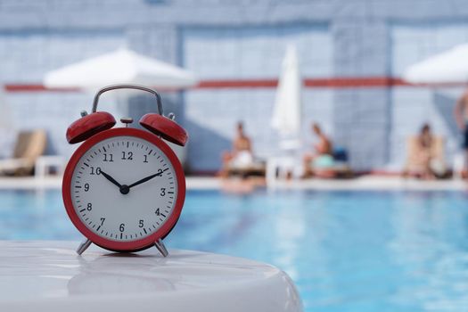 Red alarm clock standing on edge of hotel swimming pool closeup. What time can you sunbathe concept