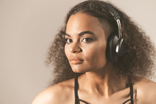 Young African-American girl listening music in headphones wearing black top isolated on grey background, emotionally move. Concept of emotions, facial expression. Toned image.