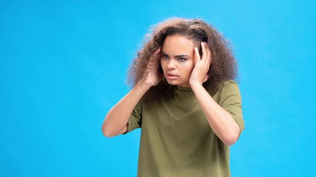 Stressed or unhappy african american girl touching her head suffering from headache wearing olive t-shirt isolated on blue background. Beauty concept. Health care concept.