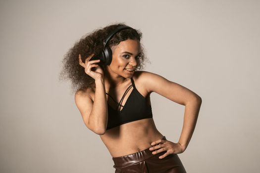 Cheerful young African-American girl listening music wearing black top and leather pants in headphones, isolated on grey background, emotionally move, have fun.
