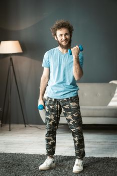 Smiling Young Man in Home Clothes does His Daily Workouts With Dumbbells at Home. Nice Man Pumps His Arm Muscles. High quality photo
