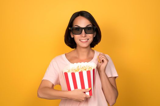 Bucket of popcorn and stereo 3D glasses in hands of a young brunette woman. Cinema concept. Isolated on yellow background.