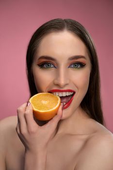 Beautiful fashion girl holding orange sliced in half in her hand biting it looking at camera. Charming joyful funny lady with red lips and long hair isolated on pink background.