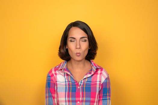 Young woman making funny faces looking to camera. Female model in plaid shirt isolated on yellow background. Human emotions, facial expression concept.